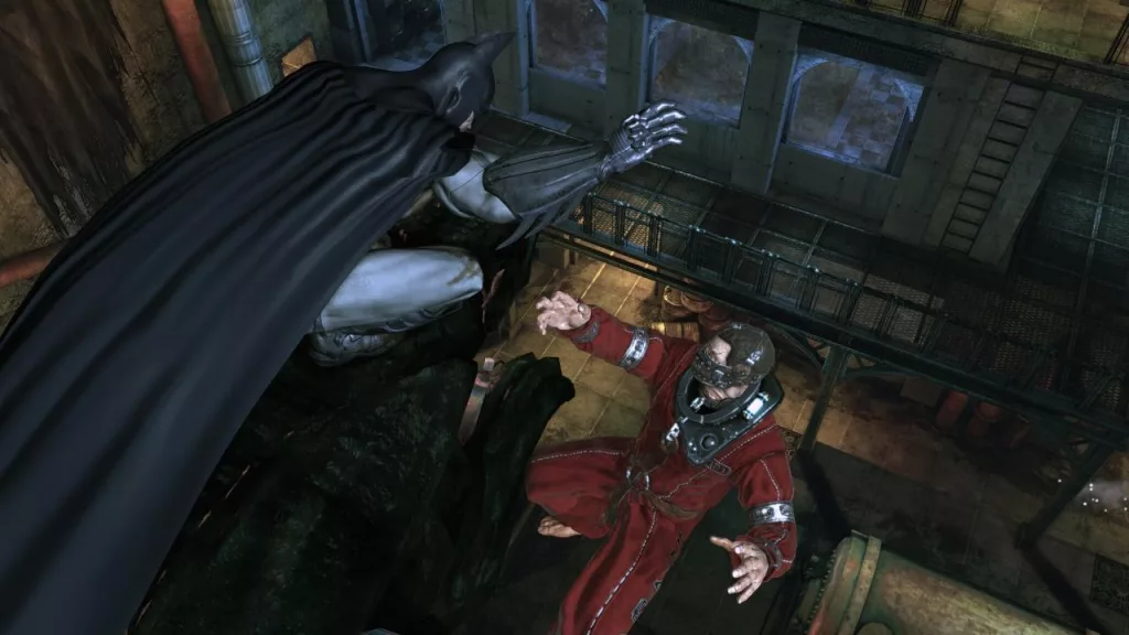 A screenshot of Batman from the video game Batman: Arkham Asylum. Batman is perched on a ledge, overlooking a room in Arkham Asylum. He is wearing his iconic black batsuit, and his cape is billowing behind him. In the background, a large, muscular inmate with a gas mask is visible.