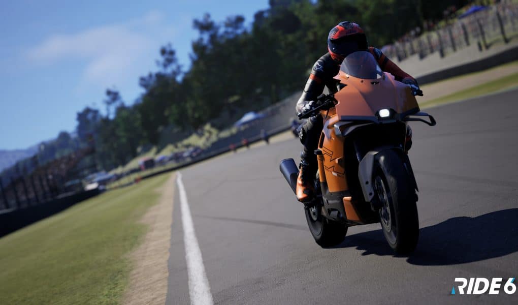 A realistic screenshot from the motorcycle racing game RIDE 6 showing a rider leaning into a turn on an orange sport bike on a paved race track with green grass and crowds in the background.