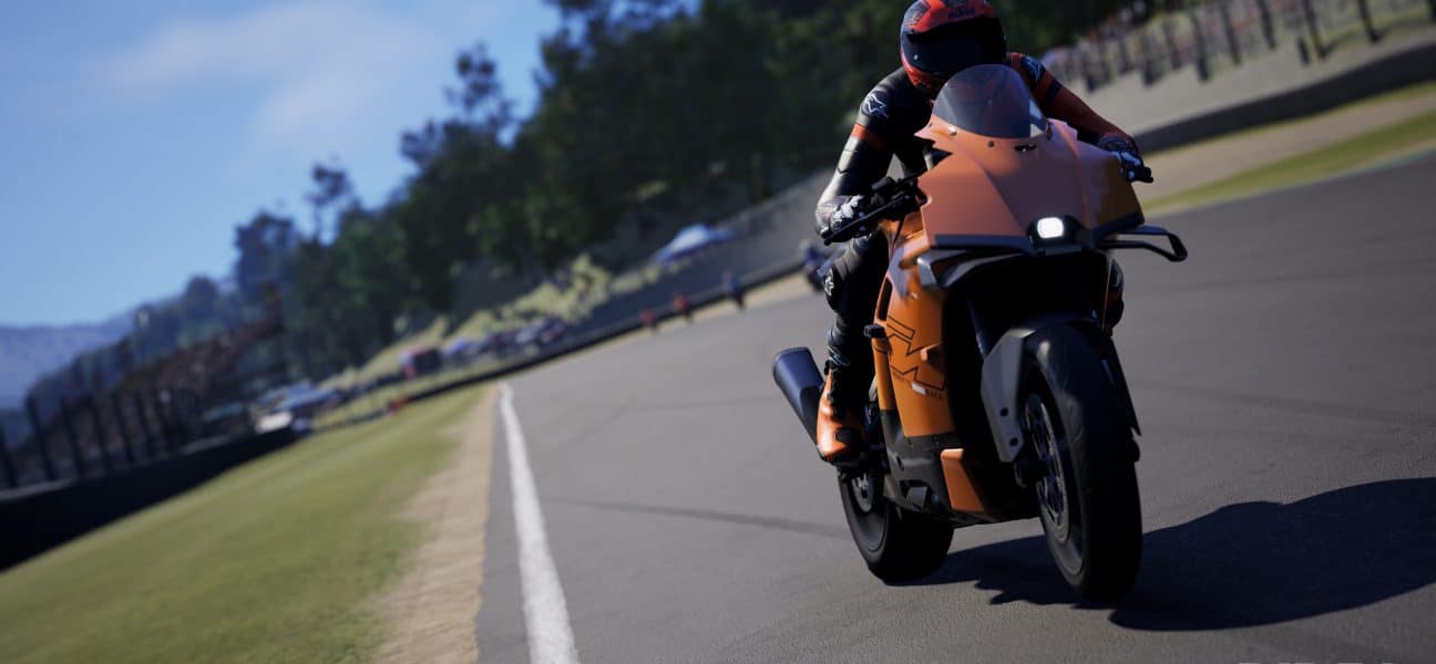 A realistic screenshot from the motorcycle racing game RIDE 6 showing a rider leaning into a turn on an orange sport bike on a paved race track with green grass and crowds in the background.