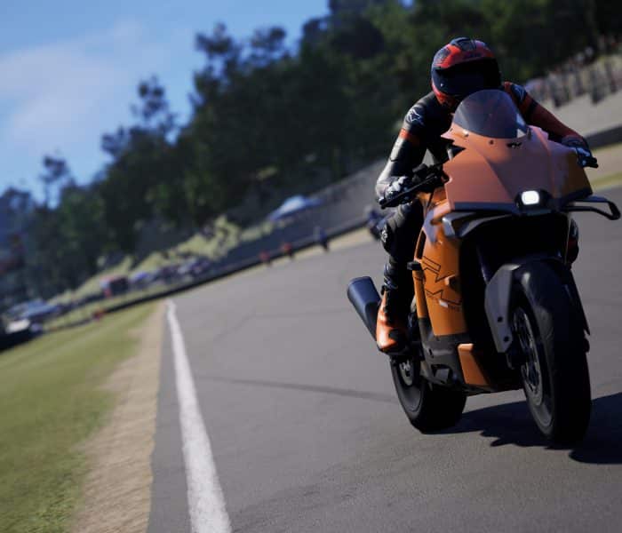 A realistic screenshot from the motorcycle racing game RIDE 6 showing a rider leaning into a turn on an orange sport bike on a paved race track with green grass and crowds in the background.