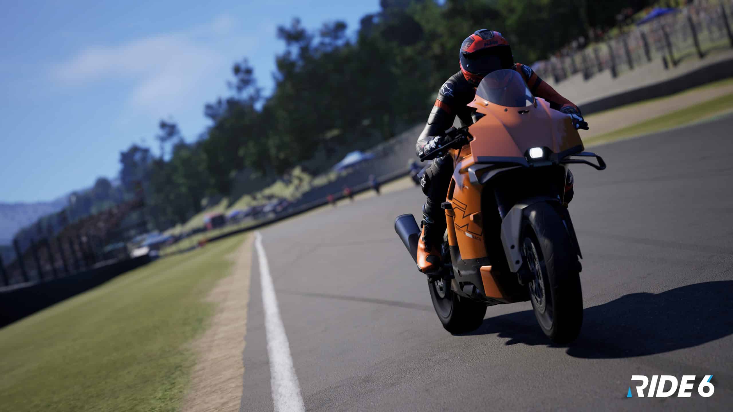 A realistic screenshot from the motorcycle racing game RIDE 6 showing a rider leaning into a turn on an orange sport bike on a paved race track with green grass and crowds in the background.