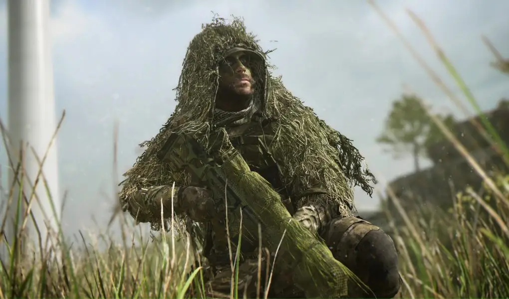 A close-up, dramatic shot of a soldier wearing a heavy ghillie suit and face paint, kneeling in tall grass with his sniper rifle, set against a blurred, slightly hazy background with a wind turbine.