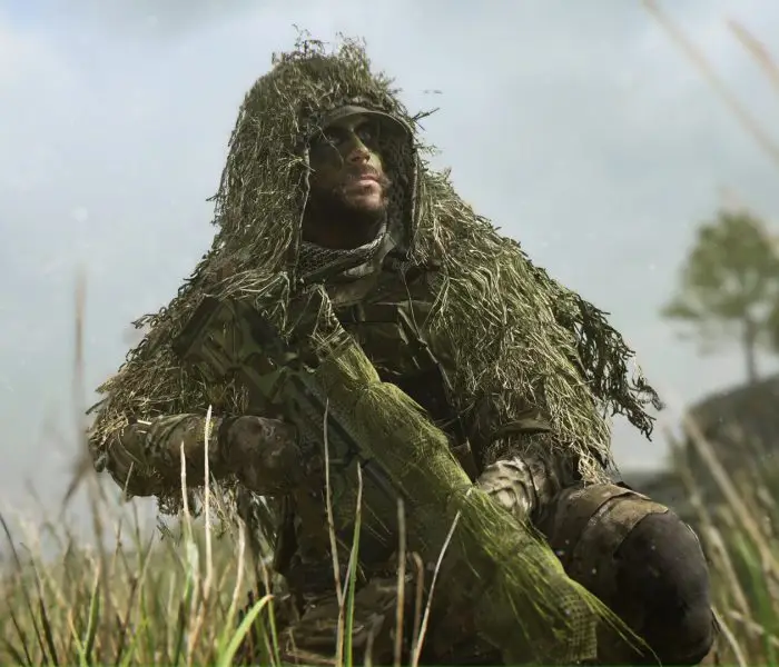 A close-up, dramatic shot of a soldier wearing a heavy ghillie suit and face paint, kneeling in tall grass with his sniper rifle, set against a blurred, slightly hazy background with a wind turbine.